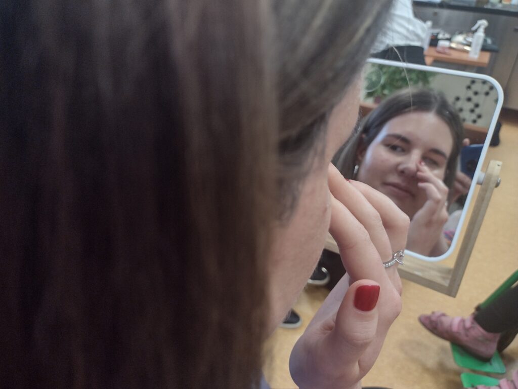 W odbiciu małego, stojącego lusterka widać uśmiechniętą twarz uczennicy, która z wyczuciem i precyzją wklepuje palcem kosmetyk w okolicę oka, samodzielnie wykonując kolejne kroki makijażu.