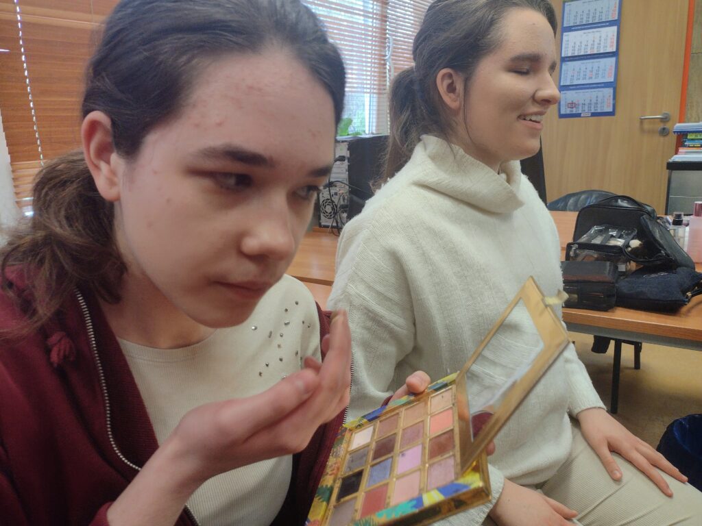 
Uczennica w bordowej bluzie z uwagą przygląda się kolorowym cieniom w złotej palecie, przygotowując się do ich nałożenia, podczas gdy siedząca obok koleżanka w białym swetrze uśmiecha się, ciesząc się wspólną nauką makijażu.