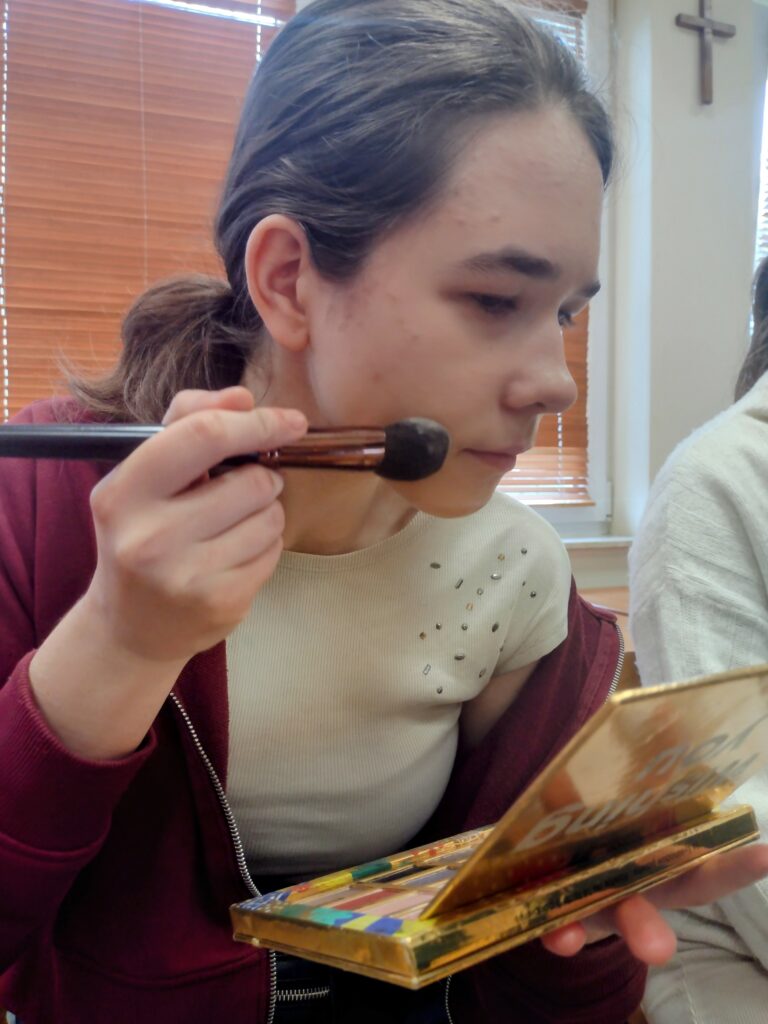 Skupiona uczennica w bordowej bluzie samodzielnie nakłada pędzlem puder na policzek, przeglądając się w lusterku złotej palety i precyzyjnie ćwicząc nowo poznane techniki makijażu.