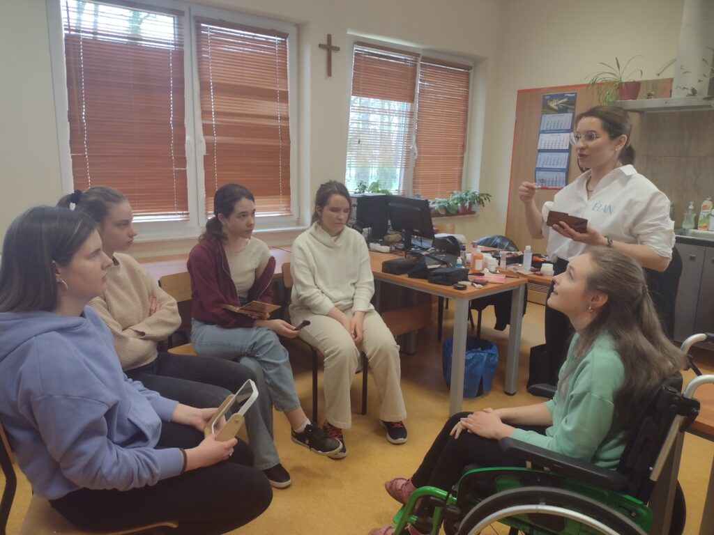 Na tle okien z brązowymi żaluzjami pięć uczennic siedzi w półkolu i z ogromnym skupieniem wsłuchuje się w rady profesjonalnej makijażystki, która stojąc przed nimi z paletą w dłoni, cierpliwie tłumaczy zasady dbania o cerę i tajniki wykonywania makijażu.