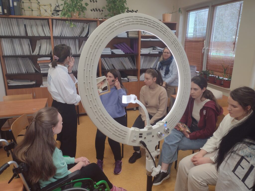 Widok przez dużą, świecącą lampę pierścieniową ukazuje grupę uczennic uważnie słuchających instrukcji makijażystki, podczas gdy jedna z nich samodzielnie nakłada kosmetyk przed małym lusterkiem, ćwicząc poznane techniki.