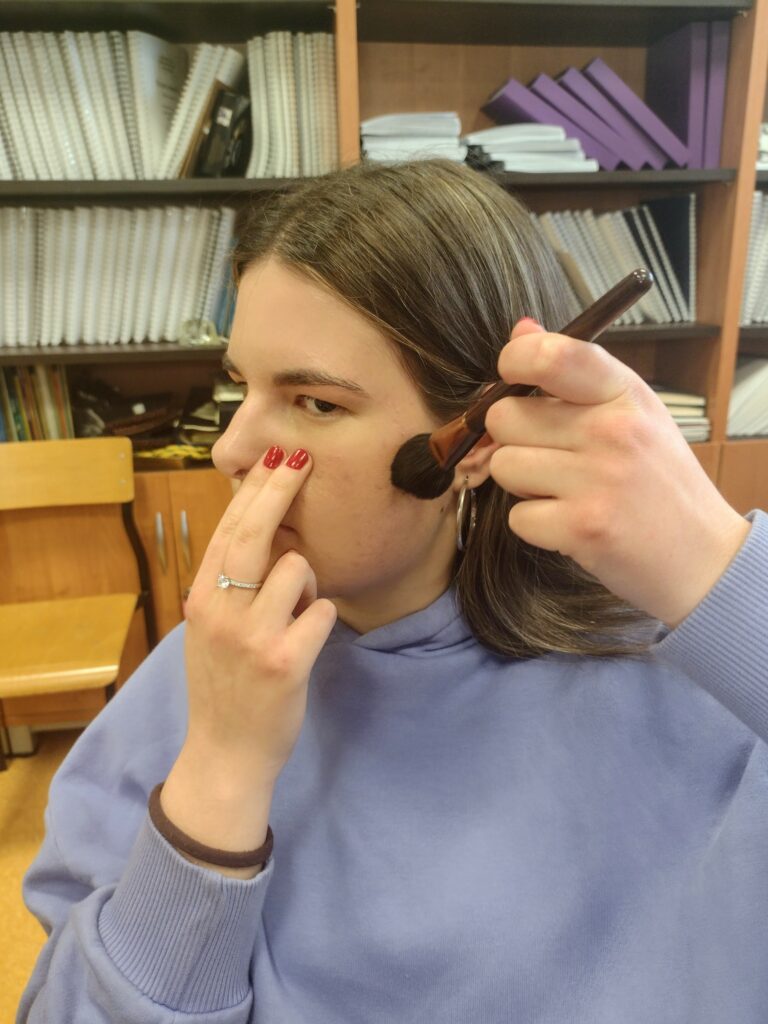 Na zdjęciu widać uczennicę w fioletowej bluzie, która z uśmiechem i dużym skupieniem samodzielnie nakłada pędzelkiem kosmetyk na policzek, wykorzystując w praktyce triki poznane podczas warsztatów makijażu.