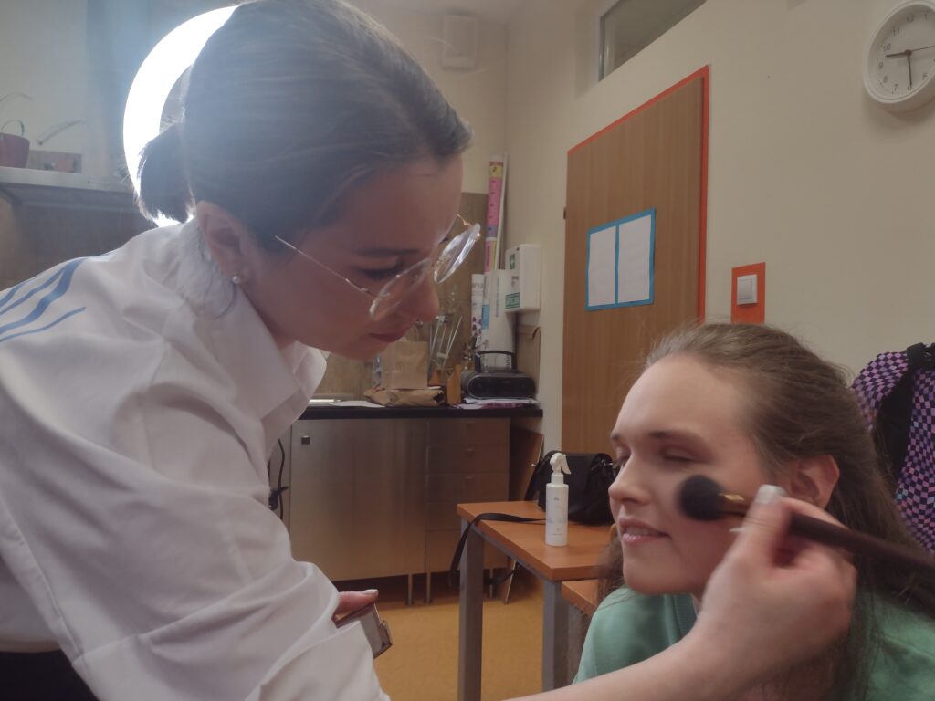 
Makijażystka w okularach, doświetlona blaskiem profesjonalnej lampy, z wyczuciem nakłada pędzlem puder na policzek uśmiechniętej uczennicy, dbając o subtelne wykończenie jej makijażu.