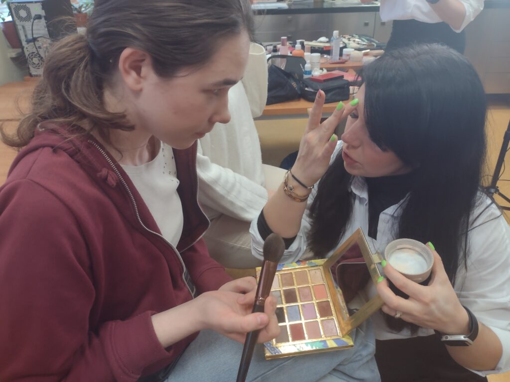 Makijażystka z uwagą instruuje uczennicę, pokazując jej na własnej twarzy technikę aplikacji, podczas gdy dziewczyna trzyma pędzel i paletę cieni, gotowa do samodzielnego powtórzenia profesjonalnego triku.