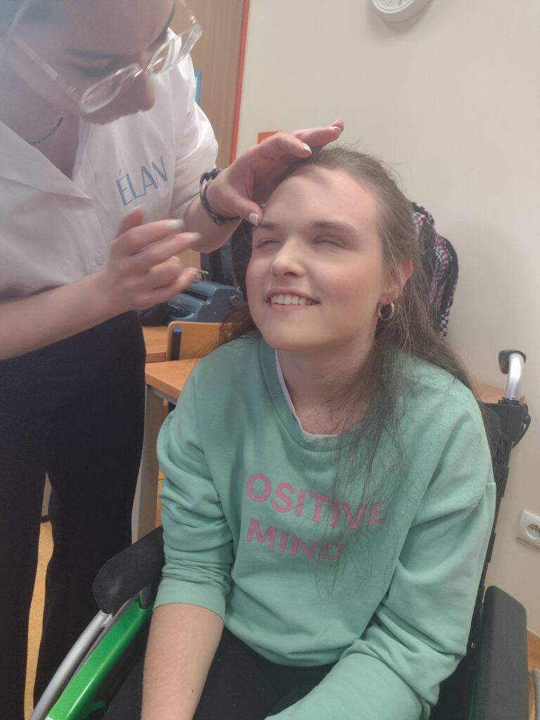 Uśmiechnięta dziewczyna w miętowej bluzie siedzi na wózku inwalidzkim, podczas gdy makijażystka w okularach delikatnie podkreśla jej brwi, tworząc radosną i pełną zaufania atmosferę podczas wspólnych warsztatów.