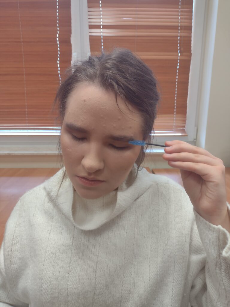 Uczennica w jasnym swetrze, z zamkniętymi oczami i pełnym skupieniem, samodzielnie wyczesuje brwi błękitną szczoteczką, ćwicząc precyzyjne wykończenie makijażu poznane podczas zajęć.