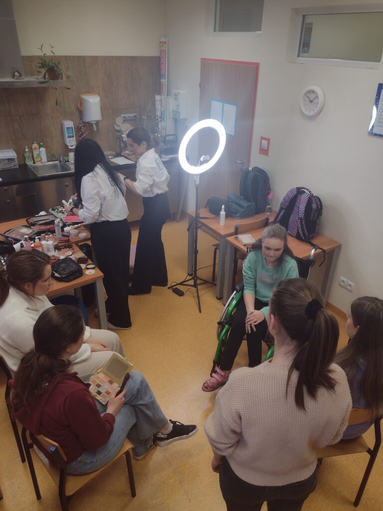 
W jasnej sali lekcyjnej, oświetlonej profesjonalną lampą pierścieniową, uczennice skupione wokół stołów pełnych kosmetyków uczestniczą w warsztatach, na których pod okiem makijażystek uczą się dobierać produkty z kolorowych palet i poznają tajniki pielęgnacji cery.