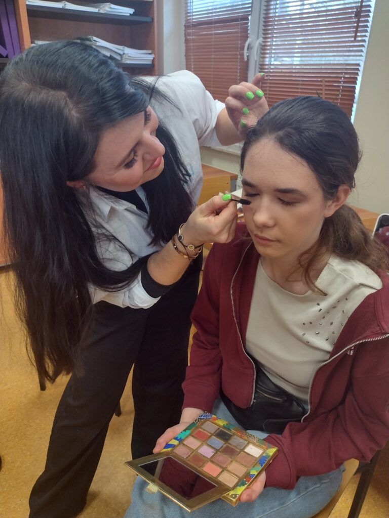 Profesjonalna makijażystka z uśmiechem pochyla się nad siedzącą uczennicą, precyzyjnie malując jej rzęsy maskarą, podczas gdy dziewczyna trzyma na kolanach otwartą paletę kolorowych cieni do powiek.