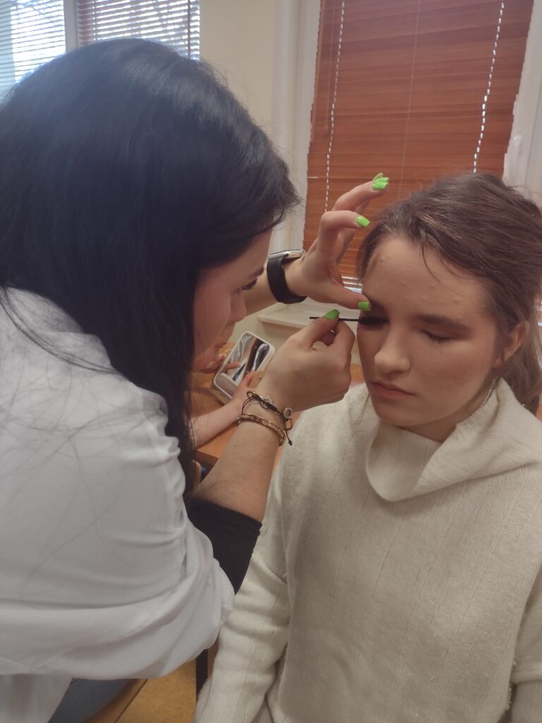 
Na zbliżeniu widać profesjonalną makijażystkę, która z dużą precyzją maluje rzęsy siedzącej, spokojnej dziewczynie, pomagając jej odkryć tajniki podkreślania urody podczas warsztatów w „Jabłonkach”.