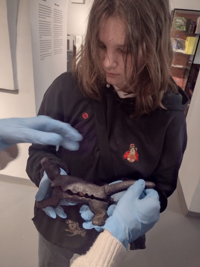 Uczennica z zaciekawieniem przygląda się zabytkowemu korkownikowi w kształcie krokodyla, odkrywając fascynujące narzędzia pracy dawnych farmaceutów podczas zwiedzania muzealnej ekspozycji.