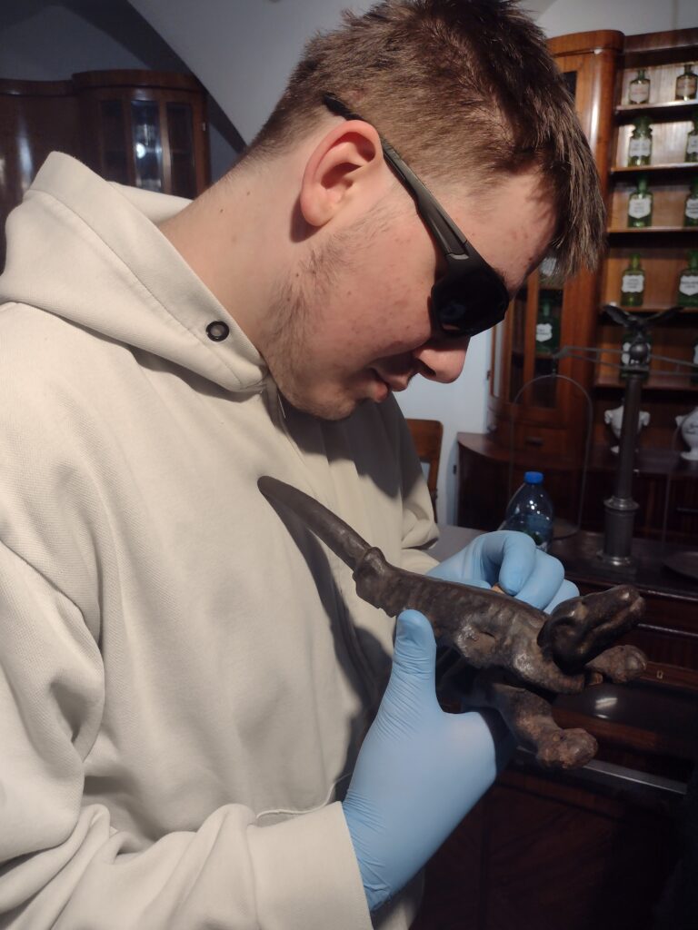 Uczeń w rękawiczkach ochronnych z zaciekawieniem ogląda nietypowy, historyczny eksponat w kształcie krokodyla, co było jedną z wielu okazji do poznania fascynujących ciekawostek z dziejów farmacji.
