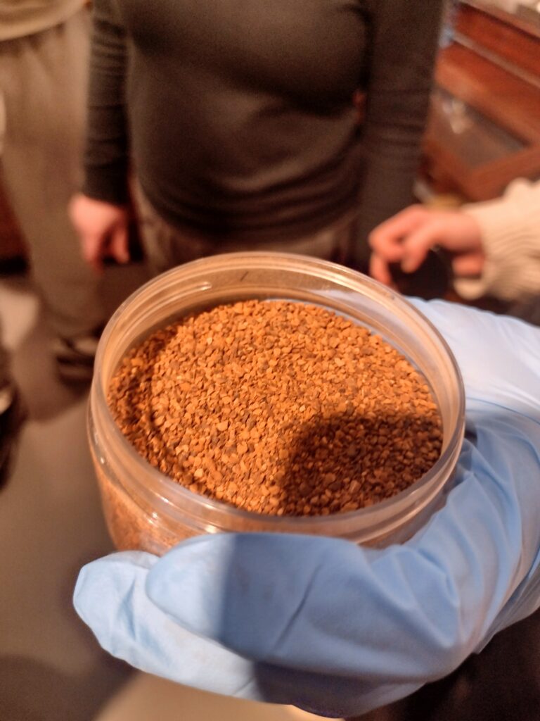 Ręka w niebieskiej rękawiczce trzyma słoik wypełniony aromatycznym granulatem, stanowiącym jeden z wielu surowców, z których uczniowie uczyli się komponować własne, unikalne wody zapachowe.