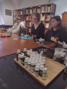 Bogatą paletę olejków eterycznych na pierwszym planie uzupełnia widok skupionych uczestników warsztatów, którzy w muzealnej bibliotece starannie komponują swoje unikalne zapachy.