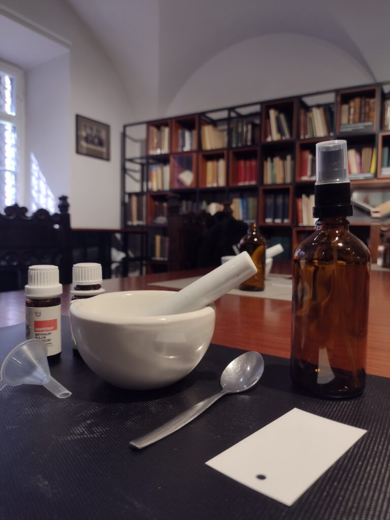 Na pierwszym planie widać zestaw do przygotowywania preparatów – porcelanowy moździerz oraz ciemną butelkę z lejkiem – podczas gdy w tle uczniowie, otoczeni regałami pełnymi książek, intensywnie pracują nad swoimi kompozycjami zapachowymi.