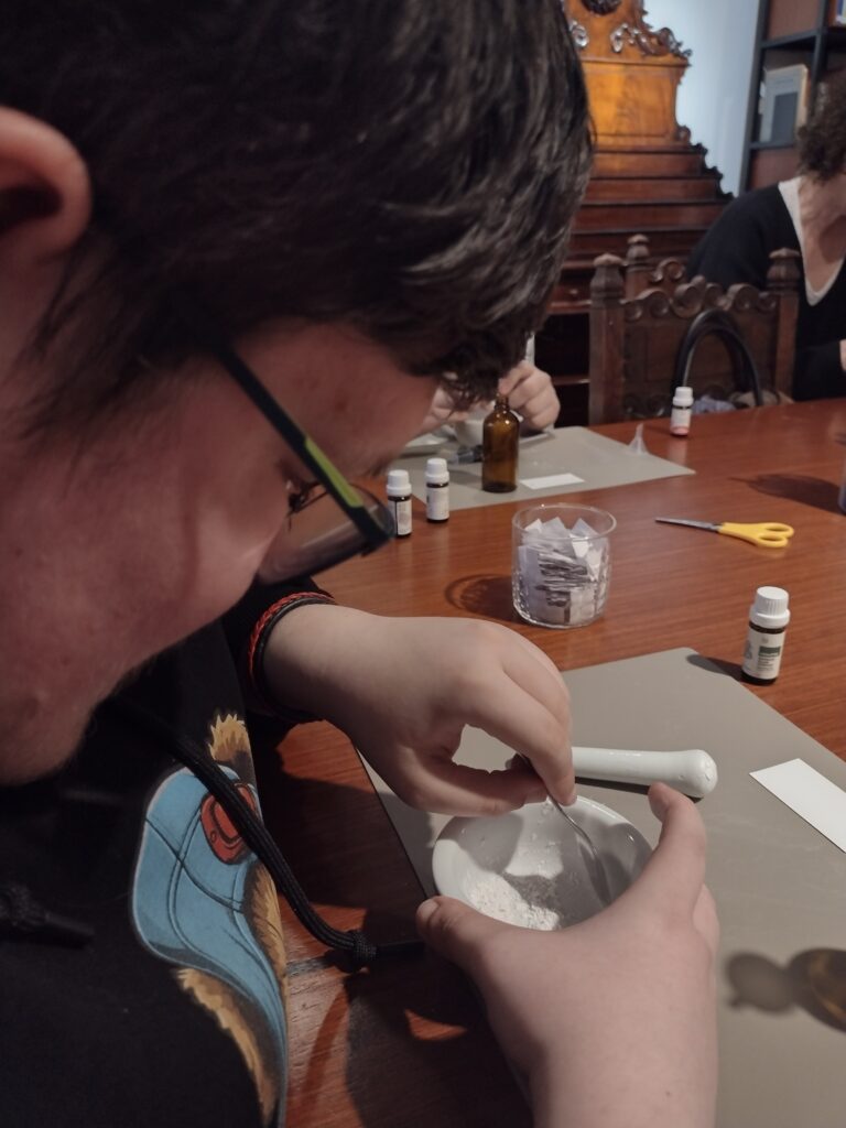 Uczeń w pełnym skupieniu uciera biały proszek w porcelanowym moździerzu, co pokazuje trud i precyzję niezbędną do stworzenia bazy pod własną, unikalną kompozycję zapachową.