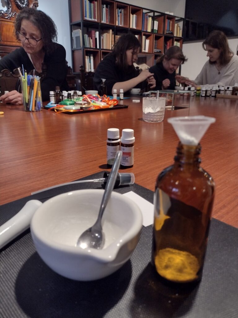 Na pierwszym planie widać zestaw do przygotowywania preparatów – porcelanowy moździerz oraz ciemną butelkę z lejkiem – podczas gdy w tle uczniowie, otoczeni regałami pełnymi książek, intensywnie pracują nad swoimi kompozycjami zapachowymi.