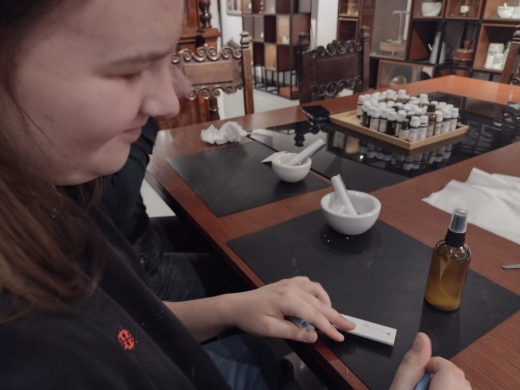 Skupiona uczestniczka warsztatów przygotowuje swoją autorską wodę zapachową przy stanowisku wyposażonym w moździerz i olejki, co idealnie ilustruje proces twórczy w zabytkowych wnętrzach Muzeum Farmacji.