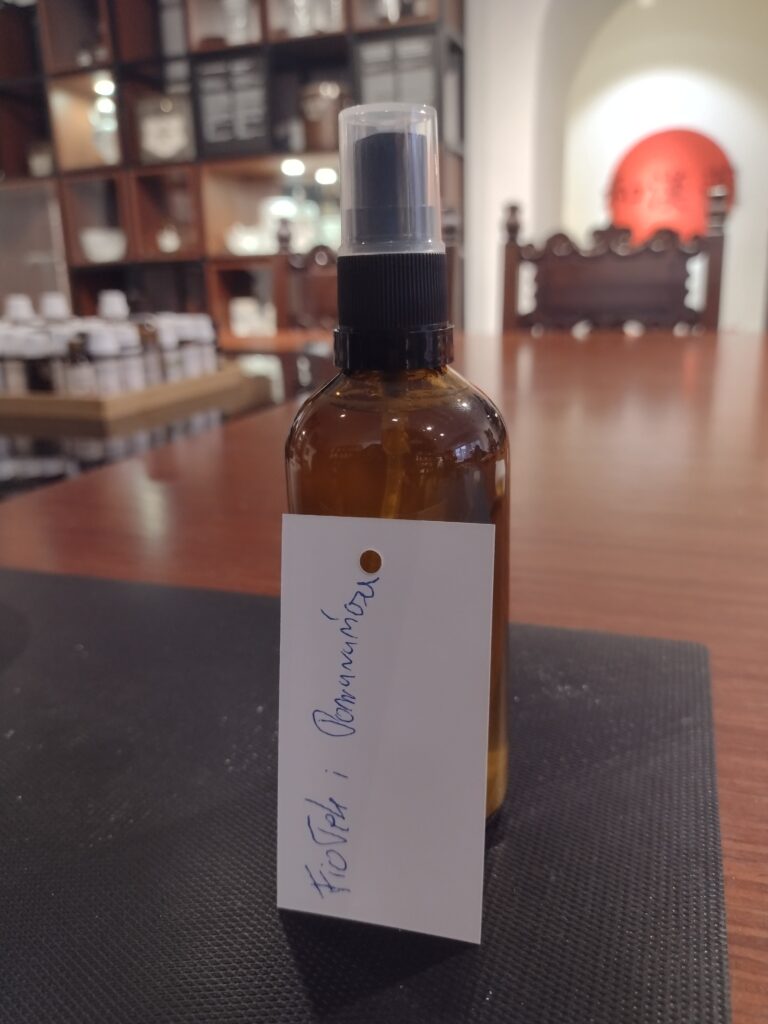 Zdjęcie przedstawia ciemną, apteczną buteleczkę z atomizerem, przy której oparta jest biała kartka z odręcznym napisem „Fiołek i Pomarańcza”, dokumentująca autorską kompozycję zapachową stworzoną przez ucznia podczas warsztatów w otoczeniu muzealnych regałów.