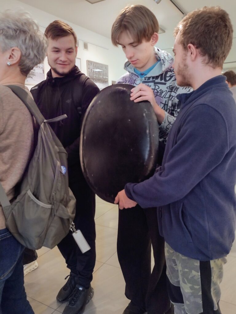 Uczestnicy warsztatów z zainteresowaniem oglądają i sprawdzają ciężar dawnej tarczy ochronnej, poznając w praktyce elementy historycznego wyposażenia Policji Państwowej.