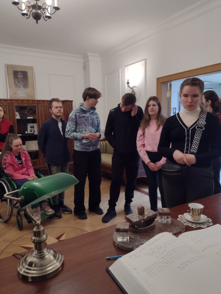 Uczestnicy wycieczki odwiedzają zrekonstruowany gabinet w Wydziale Edukacji Historycznej GKGP, gdzie z uwagą przyglądają się historycznym elementom wyposażenia biurowego, takim jak zabytkowa lampa, kałamarze oraz księgi, przenosząc się w czasie do okresu międzywojennego.