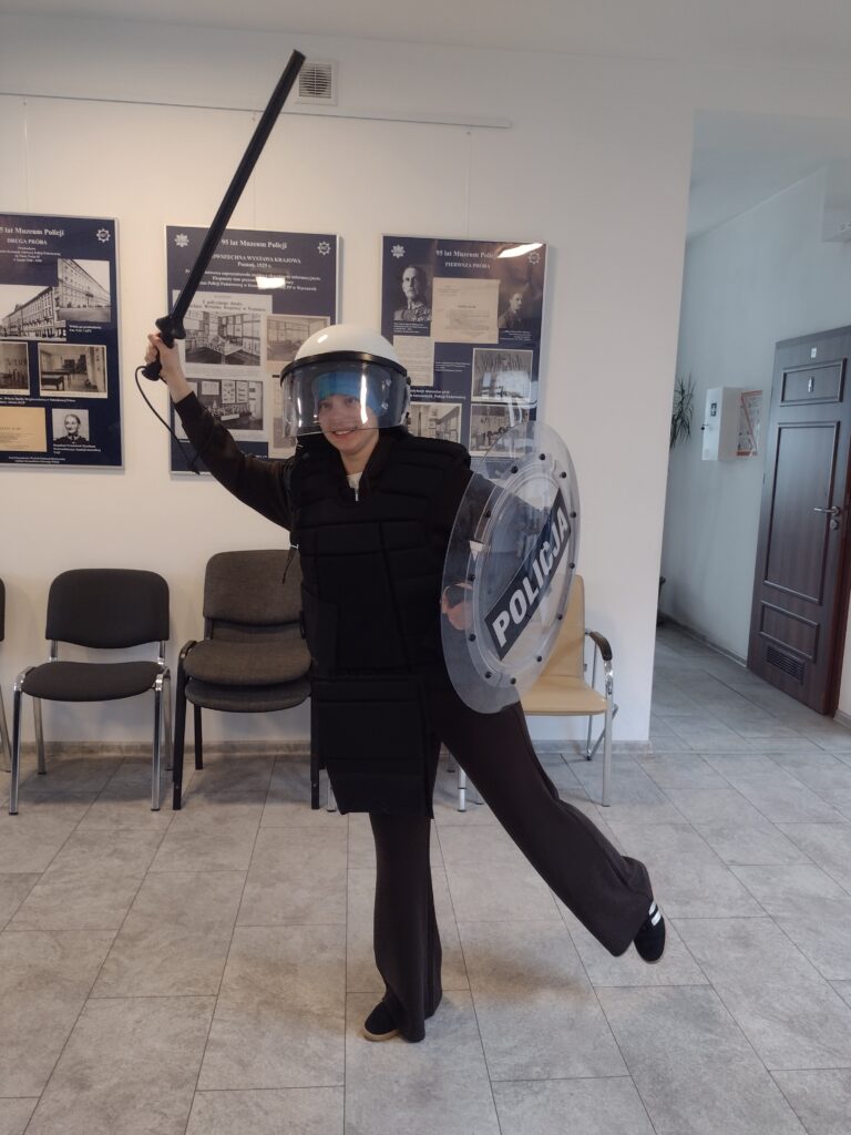 Uczestniczka wycieczki z radością przymierza współczesne elementy umundurowania, takie jak kask, kamizelka i tarcza, korzystając z atrakcji poznawania wyposażenia policyjnego w Wydziale Edukacji Historycznej GKGP.