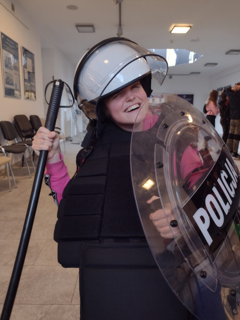 Uśmiechnięta uczestniczka wycieczki, w ramach poznawania współczesnego wyposażenia służb mundurowych, przymierza pełny rynsztunek – kask z przyłbicą, kamizelkę ochronną oraz przezroczystą tarczę z napisem „POLICJA”.