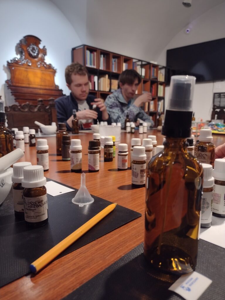 Zdjęcie przedstawia uczestników warsztatów w Muzeum Farmacji, którzy po wysłuchaniu lekcji o historii Policji Państwowej, próbują swoich sił w trudnej sztuce tworzenia własnych wód zapachowych przy użyciu licznych olejków i akcesoriów aptecznych.