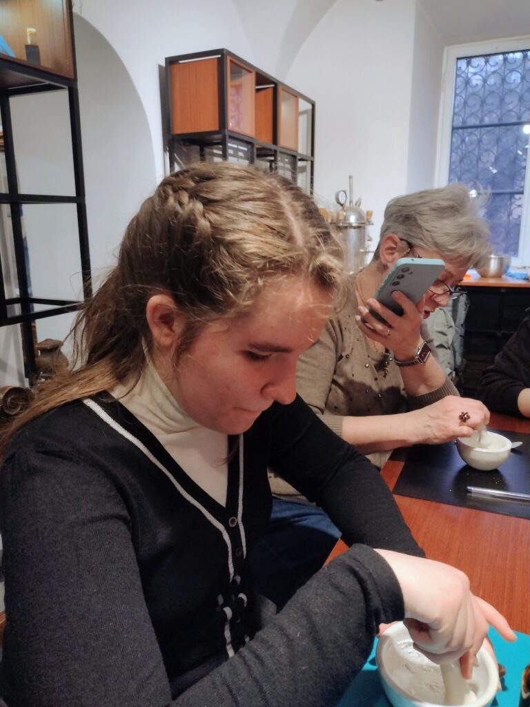 To zdjęcie przedstawia grupę osób na zajęciach w Muzeum Farmacji, gdzie przygotowują one własne wody zapachowe. Widać to po obecności moździerzy i tłuczków, które służą do rozcierania składników.