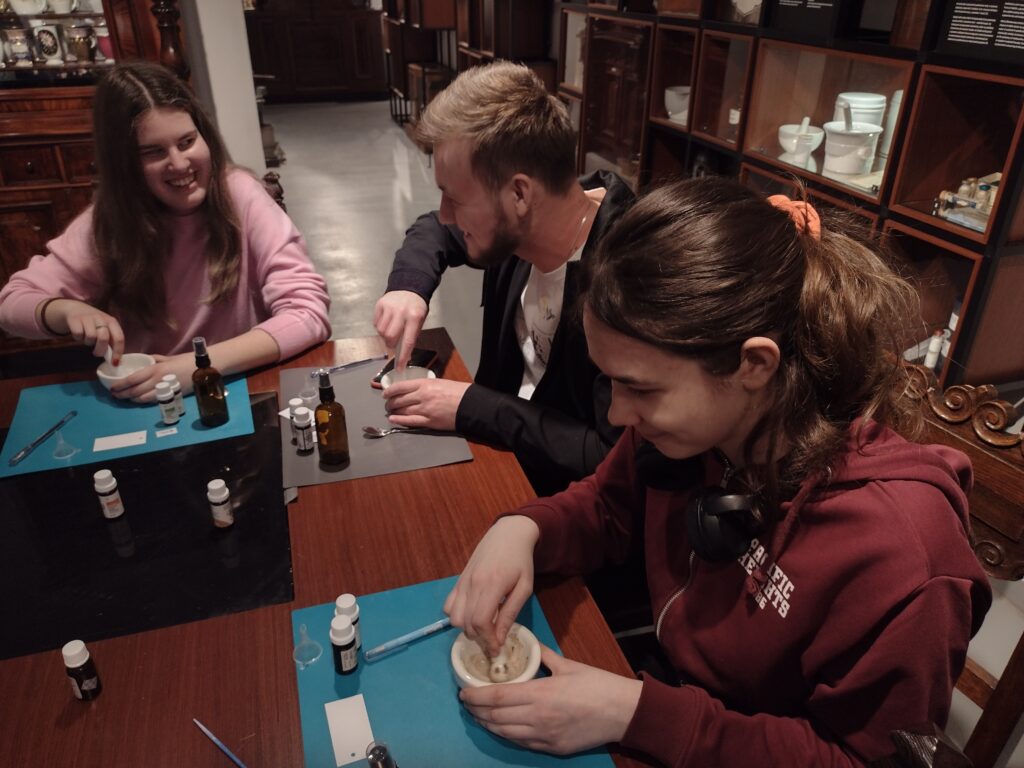 Uśmiechnięci uczestnicy warsztatów w Muzeum Farmacji z pasją ucierają składniki w moździerzach, odkrywając, że przygotowanie własnej wody zapachowej to świetna zabawa i prawdziwa sztuka wyboru.