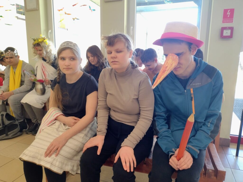 Zdjęcie przedstawia uczniów siedzących na ławce i uczestniczących w akcji „Literackie przebieranki”, wśród których widać postacie nawiązujące do bajek, takie jak Pinokio z charakterystycznym długim nosem.