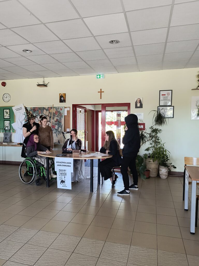 Zdjęcie przedstawia grupę przebranych uczniów zgromadzonych wokół stołu podczas teleturnieju „Książkoliada jabłonkowa”, stanowiącego główny punkt obchodów Światowego Dnia Książki w szkole.