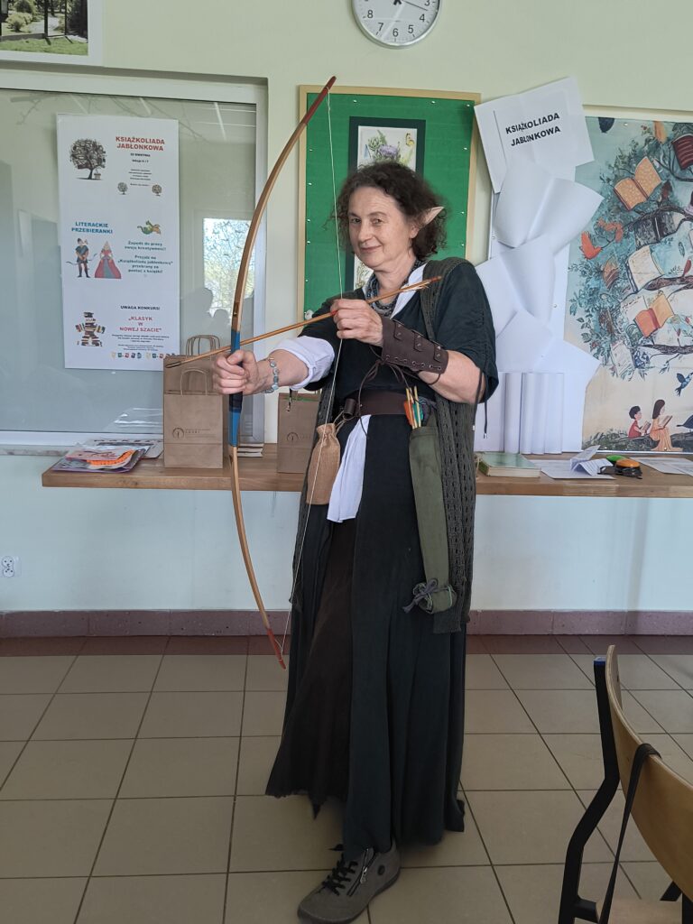 Uśmiechnięta nauczycielka w stroju elfa z łukiem i strzałą, biorąca udział w akcji „Literackie przebieranki” z okazji Światowego Dnia Książki, pozuje w szkolnym korytarzu w otoczeniu plakatów informacyjnych.