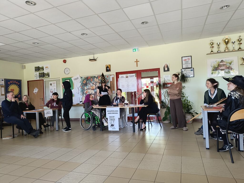 Zdjęcie przedstawia uczestników i organizatorów teleturnieju „Książkoliada jabłonkowa”, którzy zgromadzeni w szkolnej sali rywalizują w literackich wyzwaniach z okazji Światowego Dnia Książki.