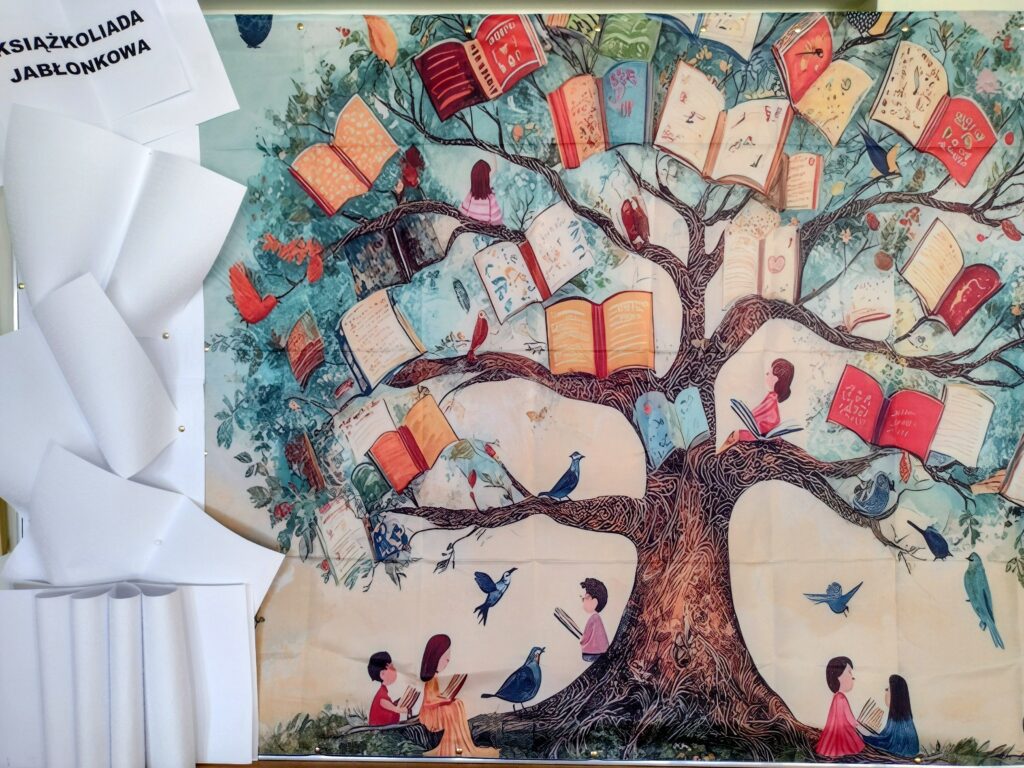 Zdjęcie przedstawia artystyczną dekorację szkolną przygotowaną na teleturniej „Książkoliada jabłonkowa”, ukazującą metaforyczne drzewo z otwartymi książkami zamiast liści, które promuje czytelnictwo wśród społeczności „Jabłonek”.