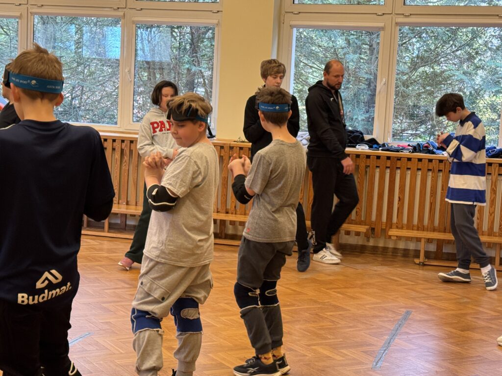 Młodzi sportowcy w pełnym skupieniu, wyposażeni w gogle startowe oraz ochraniacze na kolana i łokcie, przygotowują się do rywalizacji na boisku pod czujnym okiem instruktora.