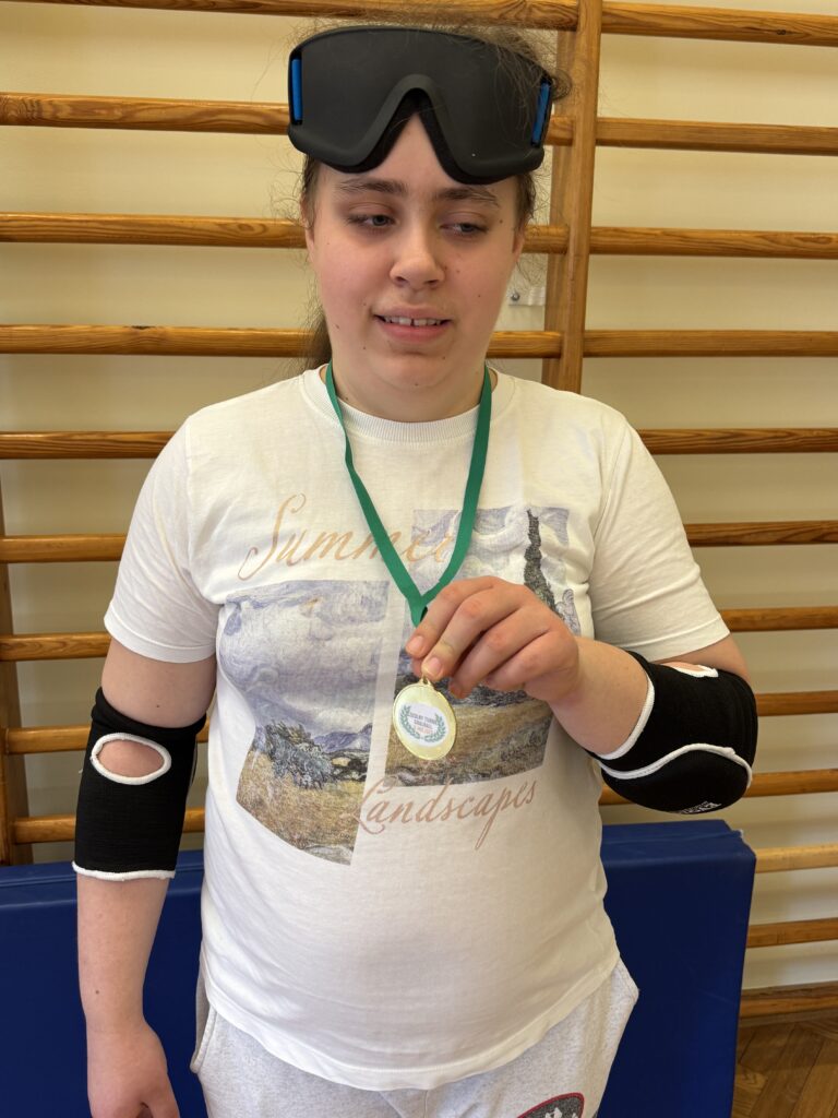 Uśmiechnięta dziewczyna w sportowym stroju i goglach na czole dumnie prezentuje zdobyty złoty medal na tle szkolnych drabinek gimnastycznych.