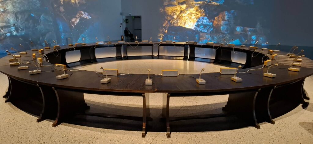 Szerokie ujęcie instalacji „Okrągły Stół. Moment przejścia” prezentuje pełną formę autentycznego mebla, który jako wystawa jednego obiektu w Muzeum Historii Polski stanowi centralny punkt refleksji nad pokojowymi przemianami demokratycznymi w naszym kraju.