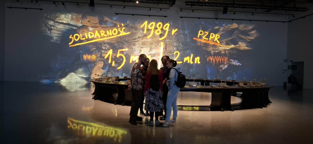 
Treść wiadomości Gemini
Grupa uczniów analizuje wystawę jednego obiektu – „Okrągły Stół. Moment przejścia”, na której multimedialna projekcja wyświetla kluczowe daty i dane historyczne z 1989 roku, stanowiąc tło dla autentycznego mebla, przy którym toczyły się obrady.