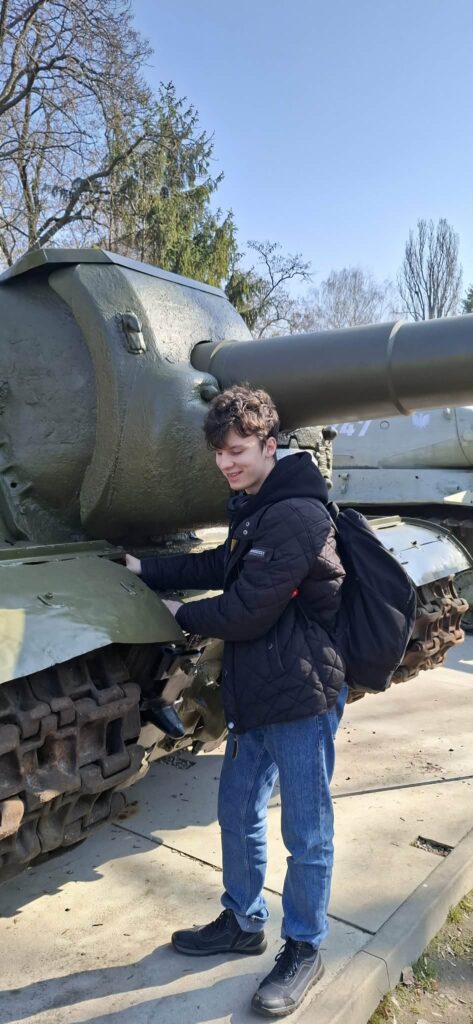 Uczeń szkoły ponadpodstawowej z ciekawością ogląda eksponaty militarne na terenie Cytadeli Warszawskiej, bezpośrednio dotykając pancerza jednego z czołgów prezentowanych na wystawie plenerowej