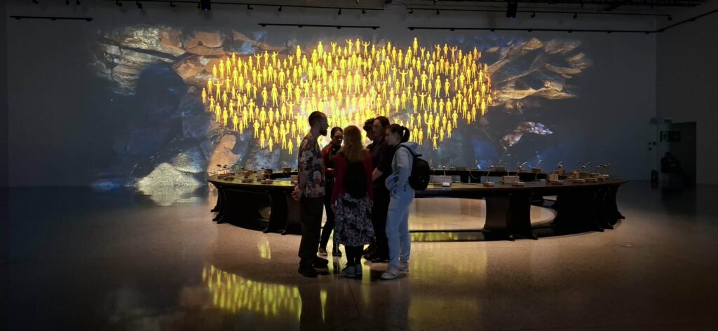 Młodzież zgromadzona wokół instalacji „Okrągły Stół. Moment przejścia” w Muzeum Historii Polski, podziwia nowoczesną formę wystawy jednego obiektu, która łączy historyczny mebel z symboliczną, świetlną projekcją tłumu w tle.