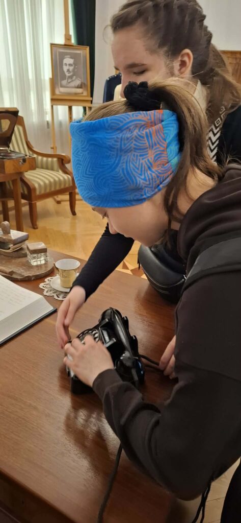 
Podczas wizyty w Wydziale Edukacji Historycznej GKGP uczestniczka z ciekawością poznaje działanie dawnego wyposażenia biurowego, oglądając z bliska zabytkowy telefon z tarczą numerową.