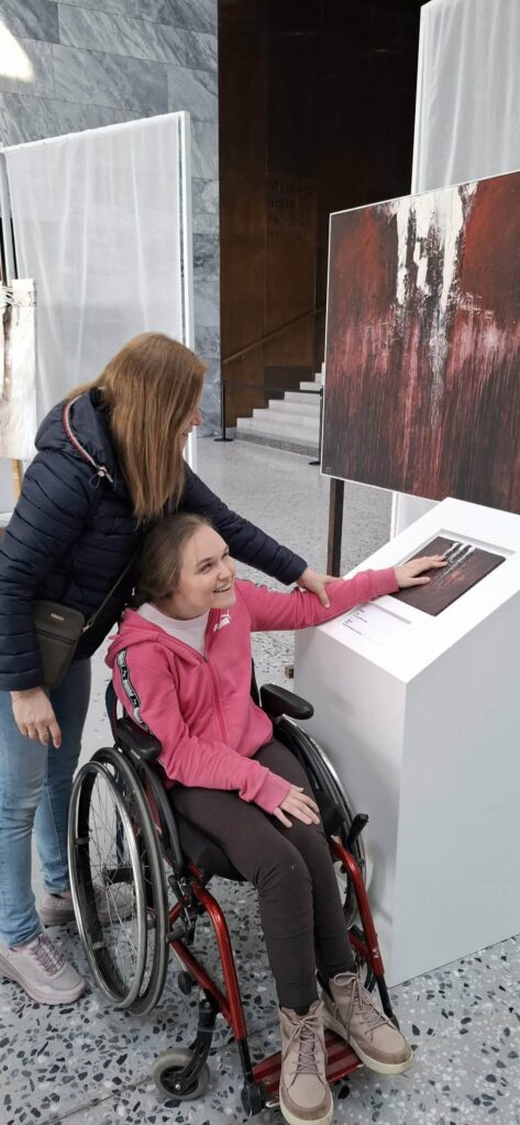
Treść wiadomości Gemini
Zdjęcie przedstawia uśmiechniętą uczennicę na wózku inwalidzkim, która wraz z opiekunką poznaje wystawę „Gero Hellmuth. Przeciw zapomnieniu” poprzez dotyk, korzystając z przygotowanych przez artystę tyflografik ułatwiających odbiór sztuki dotykiem.