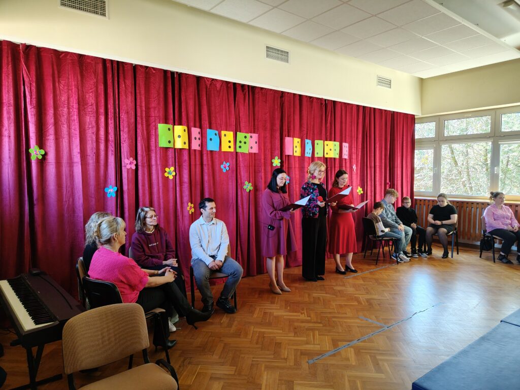 Od lewej drużyna gości, konferansjerki oraz drużyna gości na tle bordowej zasłony oraz brajlowskiego napisu Święto szkoły