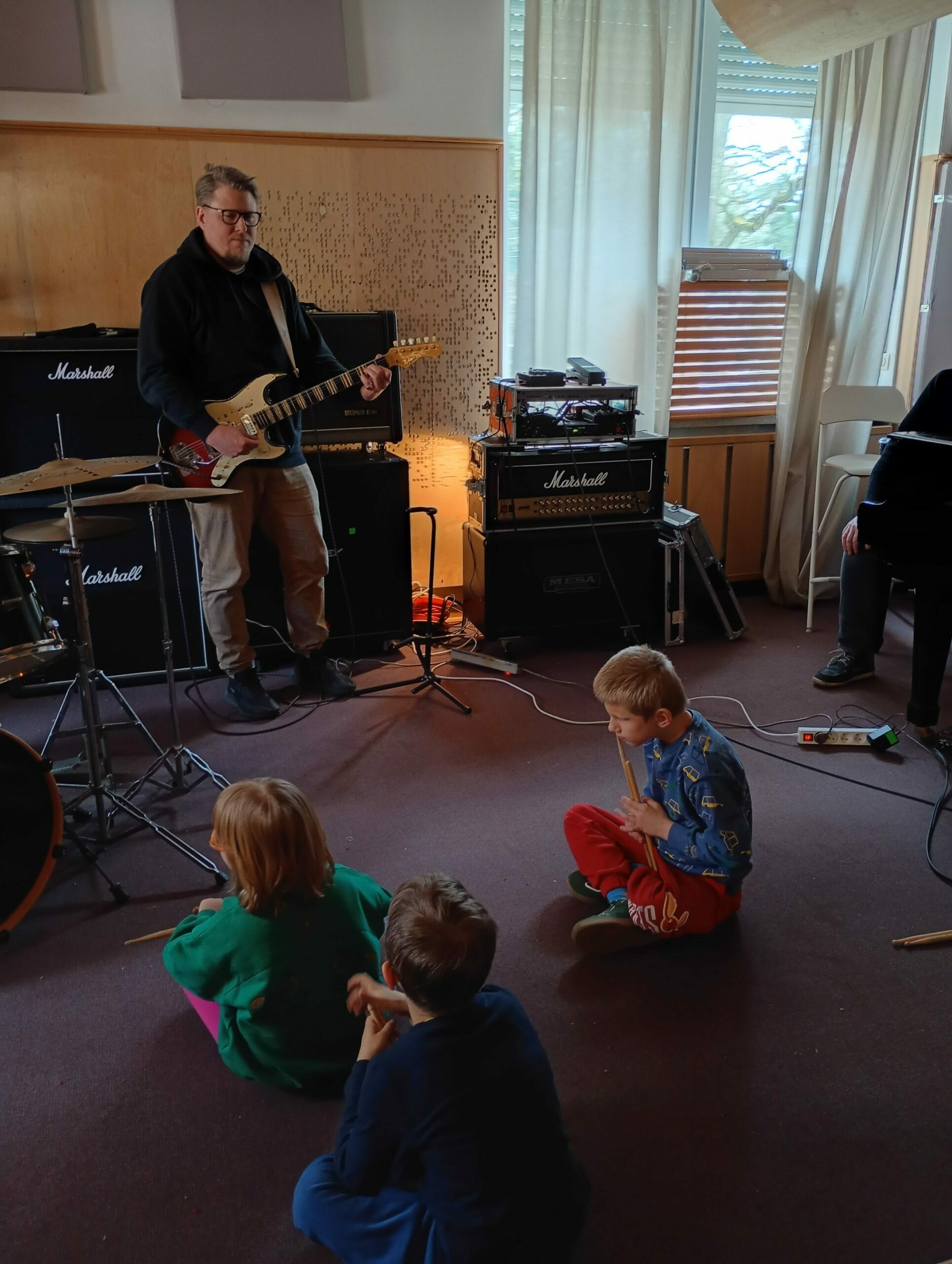 Trójka dzieci siedzi na podłodze, przed nimi  mężczyzna trzymający gitarę elektryczną.