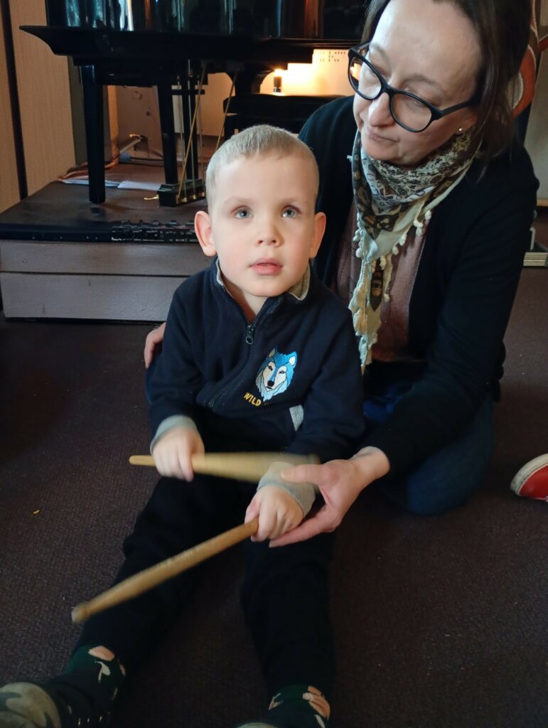 Siedzący na podłodze chłopiec trzyma w dłoniach drewniane pałeczki, za nim siedzi opiekunka,