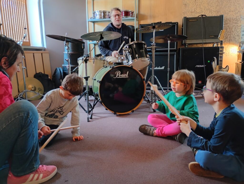 Dzieci z nauczycielką siedzą na podłodze, w dłoniach trzymają drewniane pałeczki, w głębi mężczyzna siedzący przy perkusji