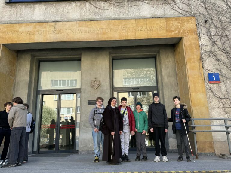 Zdjęcie przedstawia grupę uczniów klasy 8a wraz z opiekunką stojących przed wejściem do budynku Wydziału Chemii Uniwersytetu Warszawskiego przy ulicy Pasteura 1, gdzie podczas inspirującej wycieczki poznawali świat nauki, zwiedzali laboratoria i pod okiem profesjonalistów samodzielnie przeprowadzali doświadczenia chemiczne.
