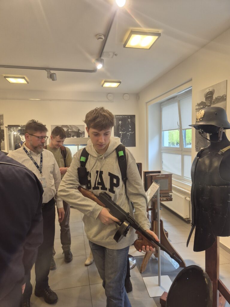 Jeden z uczniów ma okazję trzymać w rękach historyczną broń palną z bębnowym magazynkiem, oglądając dawne wyposażenie policyjne w sali wystawowej.