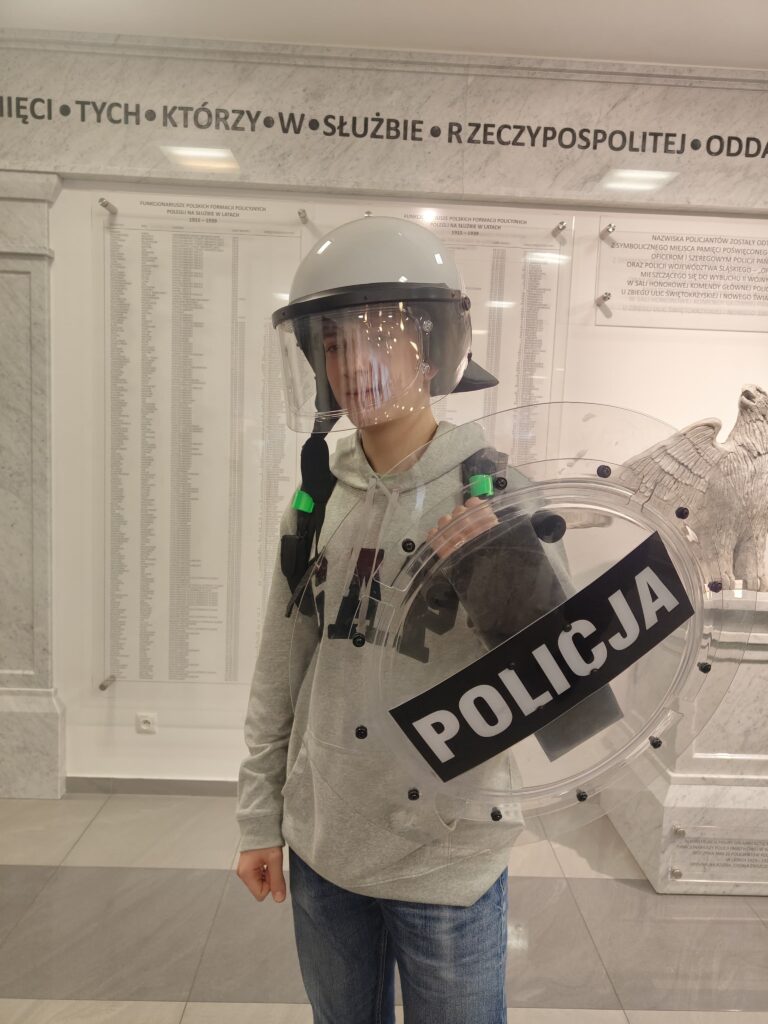 Uczeń pozuje w kasku ochronnym i z przezroczystą tarczą współczesnych Oddziałów Prewencji Policji na tle pamiątkowych tablic w Wydziale Edukacji Historycznej.