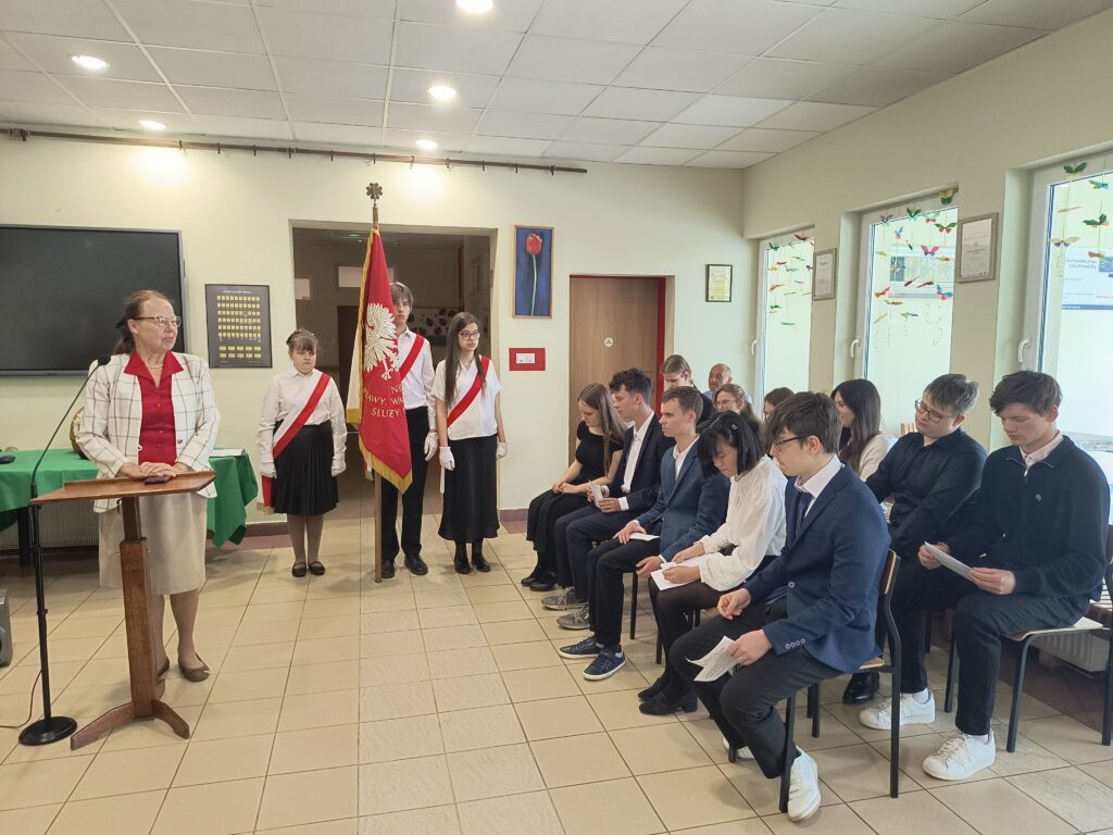 Zdjęcie przedstawia uroczystą akademię, podczas której kobieta wygłasza przemówienie przy mównicy w obecności pocztu sztandarowego oraz zgromadzonych słuchaczy.
